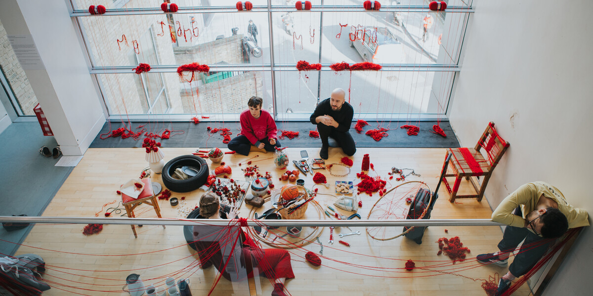 A mezzanine level floor with a wall of windows captured by an image taken slightly from above. Red yarn has been spread around the entire space, hanging over the balcony, across the widows and forming small piles on the floor. There is a large assortment of household items and trinkets that have been wrapped in red yarn. Two people sit around these items, face to face with another person, whilst a final person stands up leaning their body over the balcony.