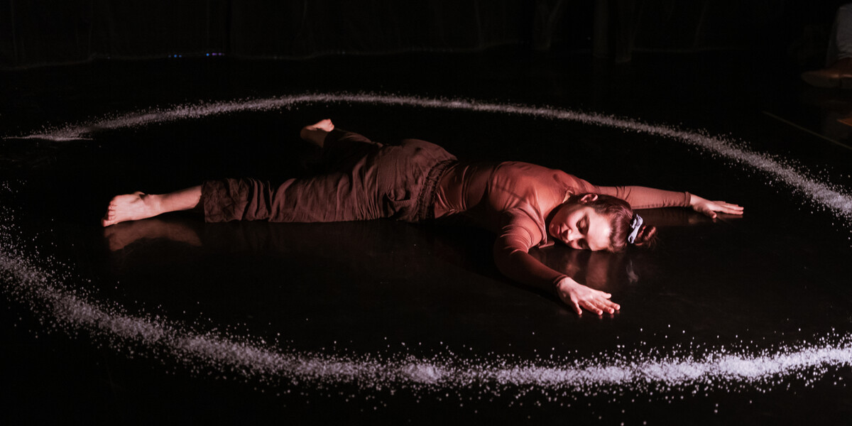 A person in a long-sleeved brown top and black trousers lies on their stomach on a dark floor, arms softly outstretched above their head and legs separated to create a star shape with their body. Around their body is a large circle of salt.