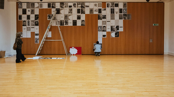 A large wooden studio and in the background you can see a wall that has A3 black and white art posters covering the majority of the wall, there is a ladder on the left hand side and a person crouching at the bottom of the wall putting up another poster