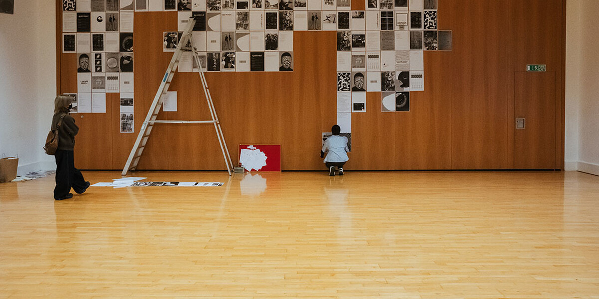 A large wooden studio and in the background you can see a wall that has A3 black and white art posters covering the majority of the wall, there is a ladder on the left hand side and a person crouching at the bottom of the wall putting up another poster