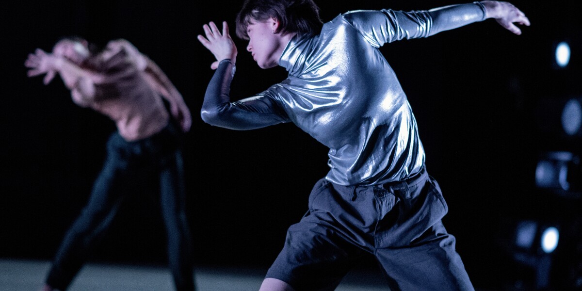 In the foreground, a dancer in black shorts and an dark shimmering turtleneck has their left arm outstretched to the side of their body, and their left bent at the elbow so their palm faces their face. They twist their body, looking over their right shoulder. Behind them is a dancer in dark trousers and a light t-shirt in a very similar position.