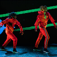 Image of two dancers in the frame. They both are wearing red tracksuits and black trainers. In the background behind them is a blue circle to the left. Two green lines also appear in the background.