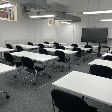 a lecture room with white desks facing a back wall. There is a TV screen on a stand on wheels at the end of the room