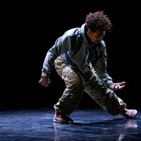 A male performer is on stage alone, he is wearing beige trousers and crouched to his right looking at his hand