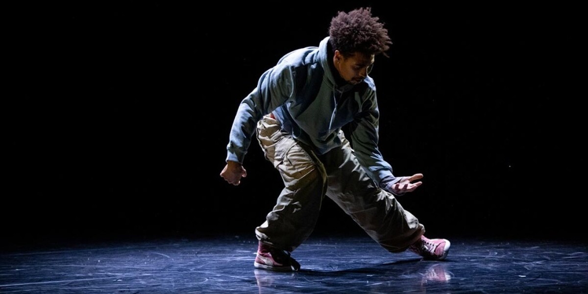 A male performer is on stage alone, he is wearing beige trousers and crouched to his right looking at his hand