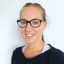 A woman with blonde hair, wearing a black long sleeved top is facing side on and smiling, she has black rimmed glassed and a white sports bra on