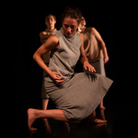 Image of a dancer dressed in a grey top and grey dress kneeing down, turning slightly to the right so their right knee is hovering off the floor. Their arms are bent slightly so their hand is by their hip.