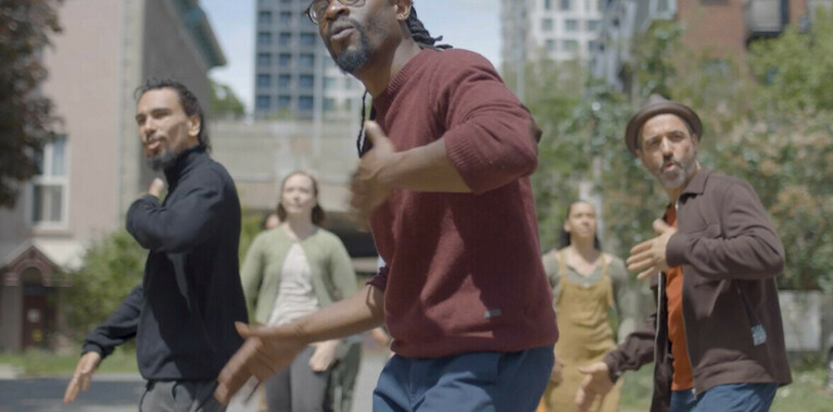 Three dancers are in a triangle formation in the foreground. They are caught in mid motion clapping their hands on their body. There is a group of dancers in two lines, slightly blurred in the background.