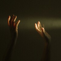 Image of two hands up in the air. A ray of light entering from the right-hand side of the image illuminates the fingers. The background is very dark.