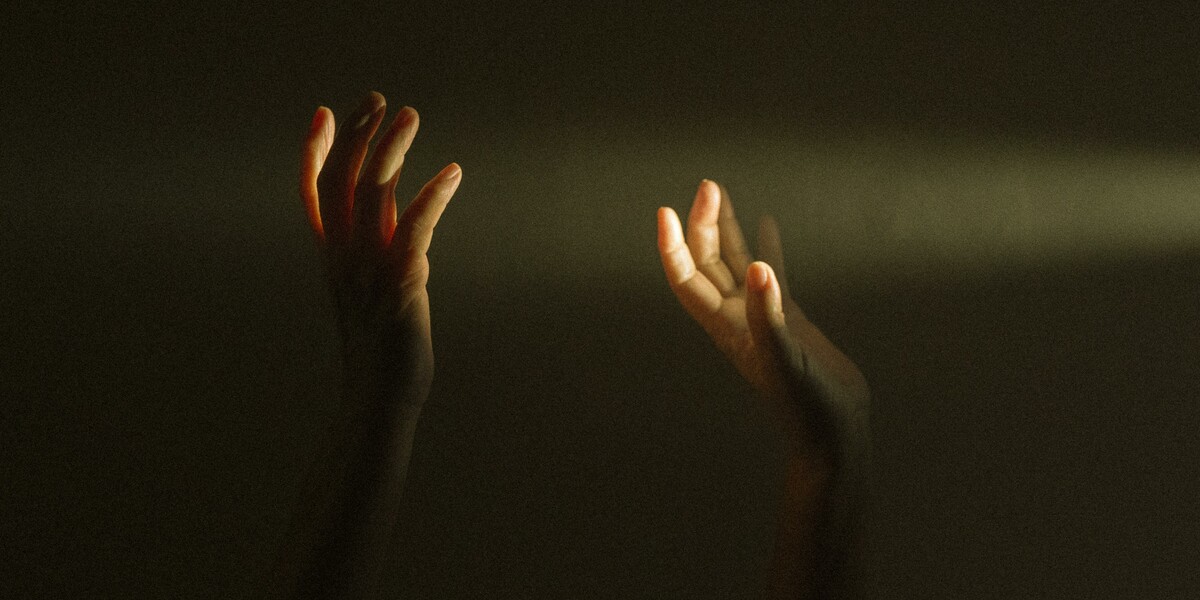 Image of two hands up in the air. A ray of light entering from the right-hand side of the image illuminates the fingers. The background is very dark.