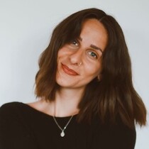A woman with shoulder length wavy hair has her head tilted slightly to the right and she is smiling at the camera. She wears a black long sleeved top with a silver chain and pendant