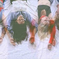 In this image four dancers lie on a white sheet with their hands above their heads. The image has been taken so that it appears the dancers are hanging upside down. Their hair is splayed on the floor and they are wearing red, black and bluish colours.