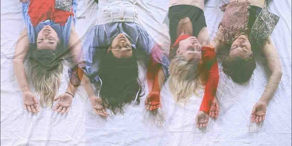 In this image four dancers lie on a white sheet with their hands above their heads. The image has been taken so that it appears the dancers are hanging upside down. Their hair is splayed on the floor and they are wearing red, black and bluish colours.