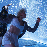 1762Three dancers are moving in front of a background where particles of light are projected. Their movements look as if they were causing the particles to movebehind them. Other elements in the space are a rocking board and knee high boots with 15cm platforms.