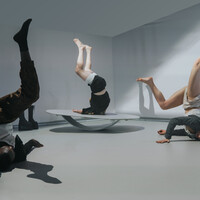 Three dancers are standing on their shoulders with their legs up against a grey background. The centre dancer is balancing on a rocking platform. Nextto it there are two male boots with a 15cm platform.