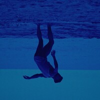 A dancer with dark-brown skin is upside down, standing in the middle of sandy terrain with water behind him. He is crouched over and dressed in a blue fish nets that cover his torso.