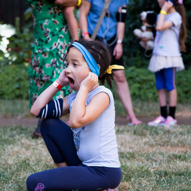 Young person sitting on grass with their fingers in their ears and their mouth open.