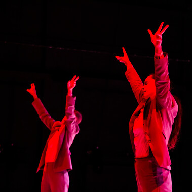 2 people on stage against a black background. They are wearing suits and raising their arms in the air. There is a red light on them.