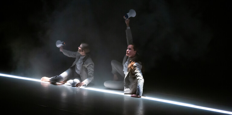 2 people sitting on stage in grey suits, the lighting dark but there is a strip of bright light