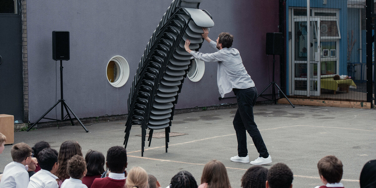 A man in a light denim shirt, dark jeans and white trainers holds a leaning tower of stacked office chairs as they lean towards him. They are stacked up higher than his head, and although they look like they might come crashing down to earth at any second, they seem suspended in the air. In the foreground is an audience of primary school students completely entranced.
