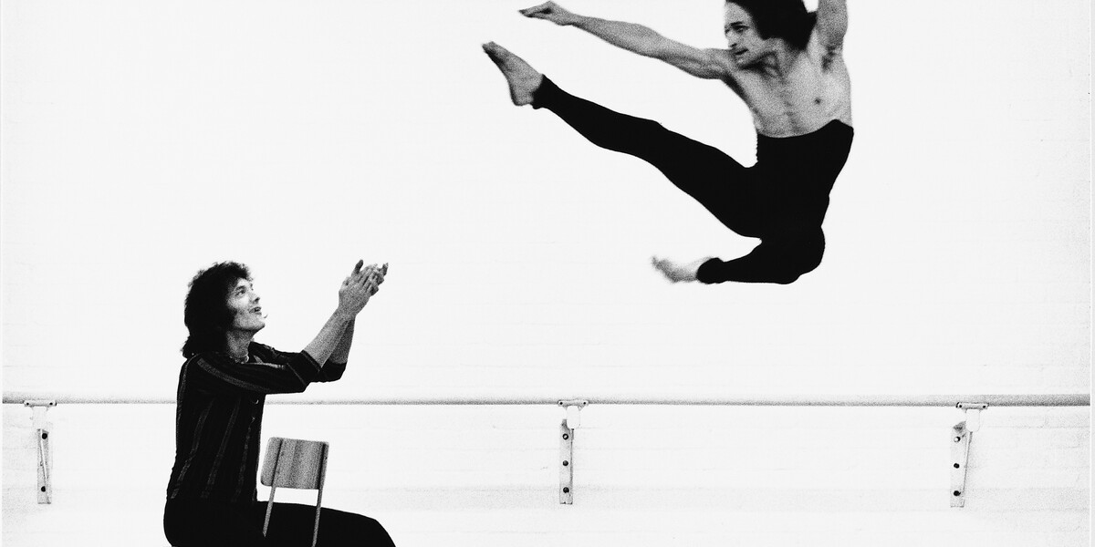 Black and White photo of Robert (Bob) Cohan in rehearsal with a member of the London Contemporary Dance Theatre. Cohan is on a chair clapping at a performer in the air with black leggings and kicking his leg forward