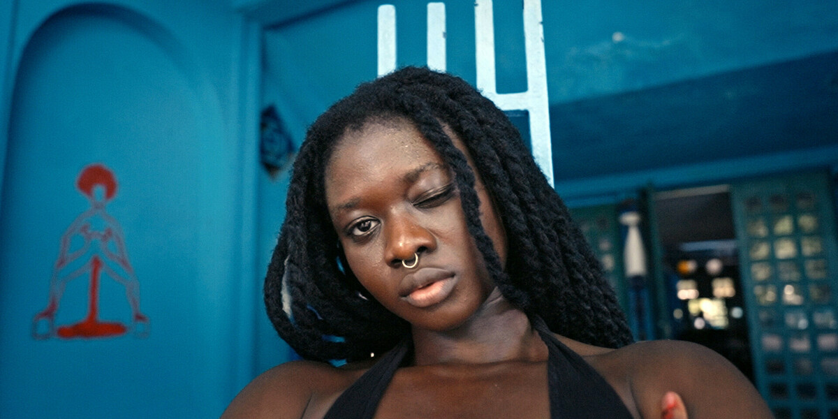 A woman is in the centre of the frame, winking into the camera. It is a close up of her face. She has long black hair and a septum piercing. The background is blue. There is wall art to the left of the woman showing a woman standing with her legs apart, blood flowing from between her legs.