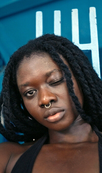 A woman is in the centre of the frame, winking into the camera. It is a close up of her face. She has long black hair and a septum piercing. The background is blue. There is wall art to the left of the woman showing a woman standing with her legs apart, blood flowing from between her legs.
