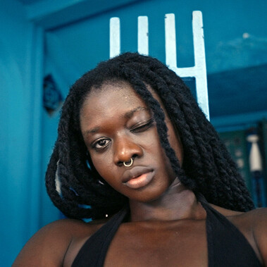 A woman is in the centre of the frame, winking into the camera. It is a close up of her face. She has long black hair and a septum piercing. The background is blue. There is wall art to the left of the woman showing a woman standing with her legs apart, blood flowing from between her legs.