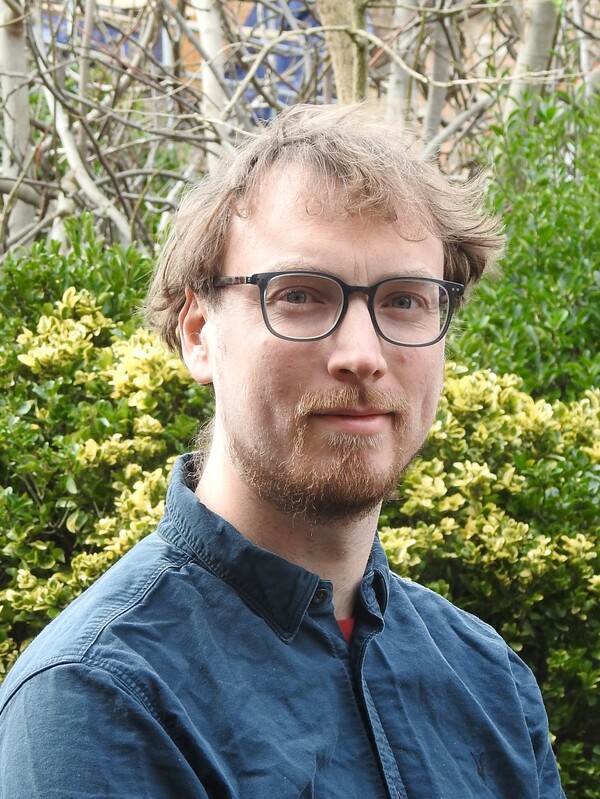 A man wearing a dark blue shirt and blue framed glasses looks into the camera. In the background there are green bushes and trees, he has light brown hair and a beard