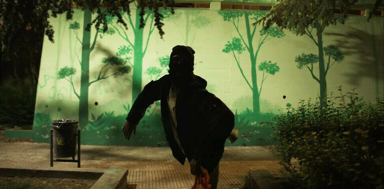 Set in a dark park, the image is of a man with a mask on, moving his body to the right. The background is a wall with painted trees and there is a rubbish bin to the left and a bush to the right.
