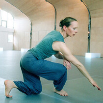 A woman wearing shades of green and blue is in a low lunge with bent knees. Her right arm is outstretched to the diagonal corner and her eyes are looking along that arm. The studio she is in curved ceilings and is name from wood.