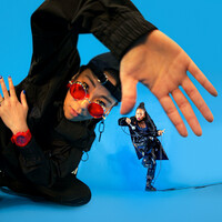 A dancer in all black trackies, a backwards black cap and red sunglasses and watch leads into the camera, their hand is up to the camera and is warped by a fish eye lense. In the background, another dancer in all black dances with a microphone in their hand. They slot into the space between the thumb and forefinger of the dancer in the foreground. The background is a bright electric blue.