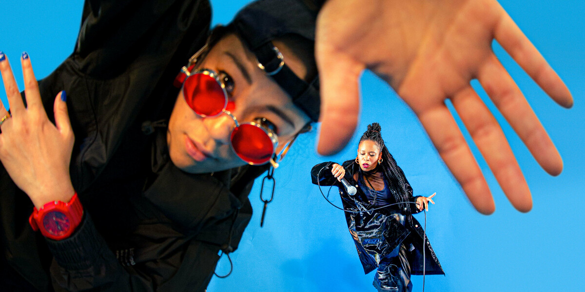 A dancer in all black trackies, a backwards black cap and red sunglasses and watch leads into the camera, their hand is up to the camera and is warped by a fish eye lense. In the background, another dancer in all black dances with a microphone in their hand. They slot into the space between the thumb and forefinger of the dancer in the foreground. The background is a bright electric blue.