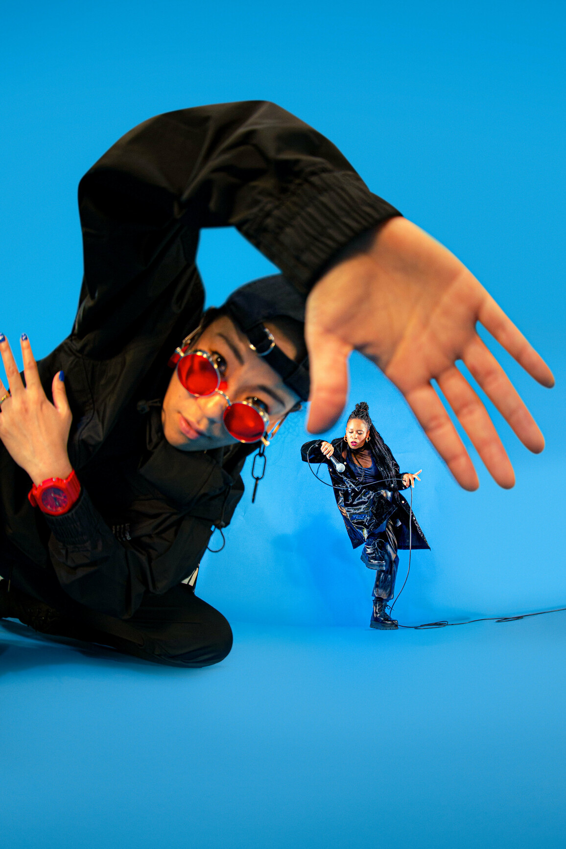 A dancer in all black trackies, a backwards black cap and red sunglasses and watch leads into the camera, their hand is up to the camera and is warped by a fish eye lense. In the background, another dancer in all black dances with a microphone in their hand. They slot into the space between the thumb and forefinger of the dancer in the foreground. The background is a bright electric blue.