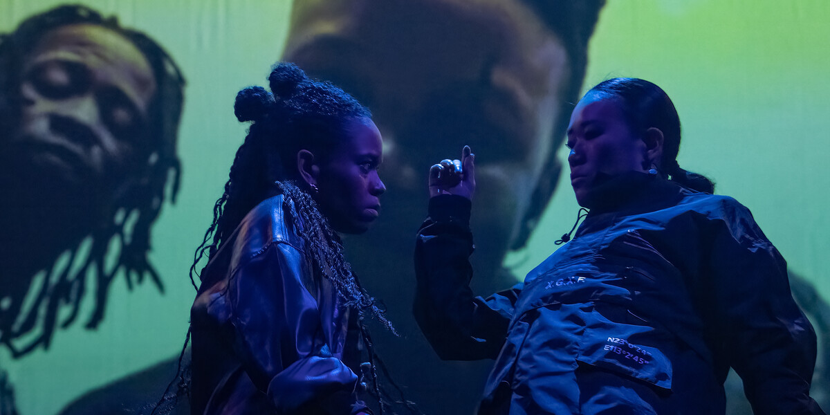 Yuki (who is East Asian, Japanese) and Sarah (who is mixed race, white British and west African, with long dark brown twists) groove intensely together, wearing matching black shiny outfits. In mid-motion, Yuki sways backwards to the left with one arm raised in the middle of the image, while Sarah leans towards them, gazing intently. Behind them, a music video is being projected with three singers looking down on them.