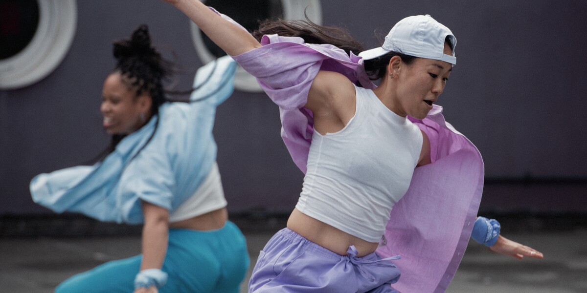 Two performers are seen dancing outside. One is wearing pastel purple shirt and trousers with a white vert and a white cap, the other, behind her is wearing matching pastel blue shirt and trousers and a vest