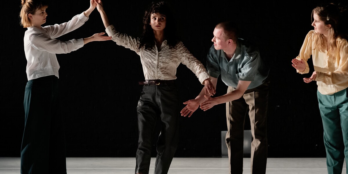 Four dancers lit by a white light on stage wear shirts and trousers in neutral colours. One dancer faces forwards but their eyes look towards stage left. Their right leg is stretched in front of them with the toe pointed and touching the floor. Their left arm is down, and their right arm reaches upwards, softly bent at the elbow. The dancer to their right holds their elbow with the tips of their fingers from underneath, and is also holding onto their right hand. To the right of them, two dancers lean their upper bodies forward and twist towards the first two, their arms in front and palms facing together mid-clap.