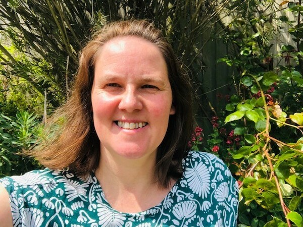 A woman with mid length brown hair is smiling and looking at the camera. She is wearing a green and white floral top and behind her there are plants and flowers.