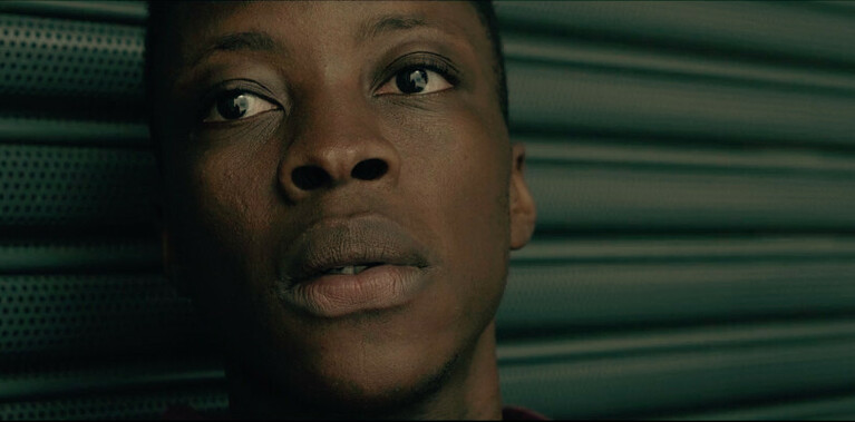 Close-up of a young man’s face, his eyes are looking to the side. The background is composed of dark grey metallic shutters.