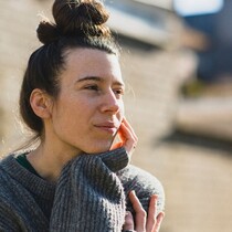 a portrait image of Simone outside, she has long brown hair tied in a bun on the top of her head and is wearing a grey woollen jumper. She is looking out of the frame toward the right.