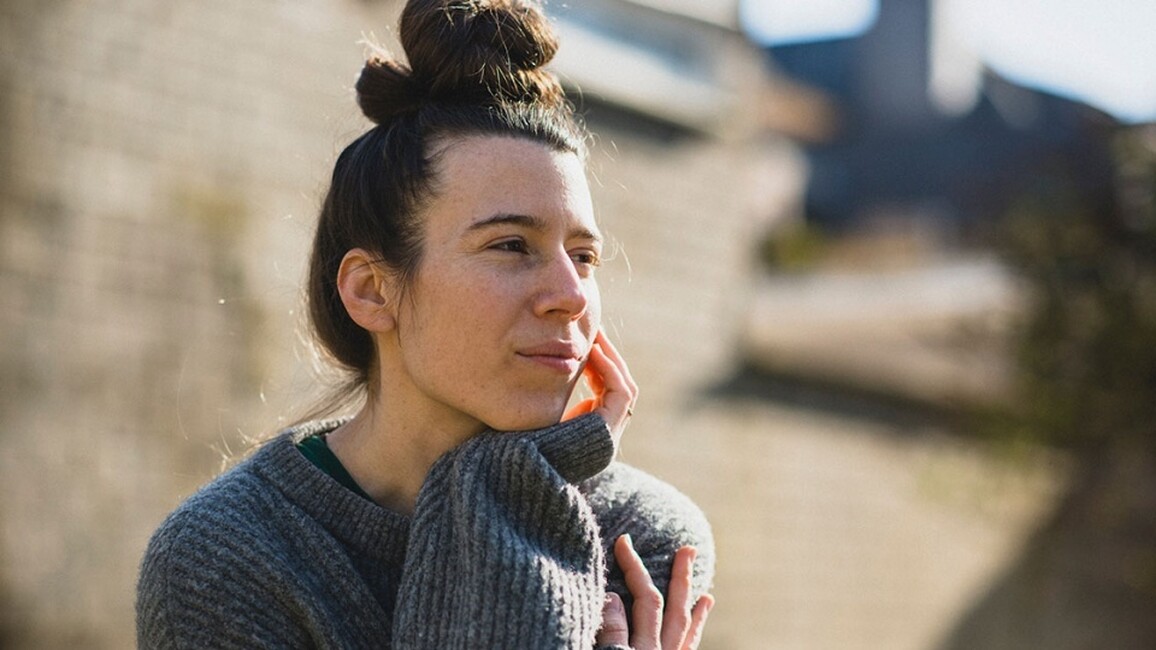 a portrait image of Simone outside, she has long brown hair tied in a bun on the top of her head and is wearing a grey woollen jumper. She is looking out of the frame toward the right.