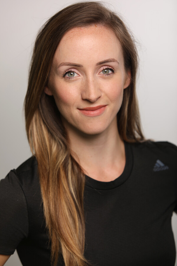 A woman with long light brown hair, wearing a black short sleeved top smiles and looks into the camera, there is a white background behind her