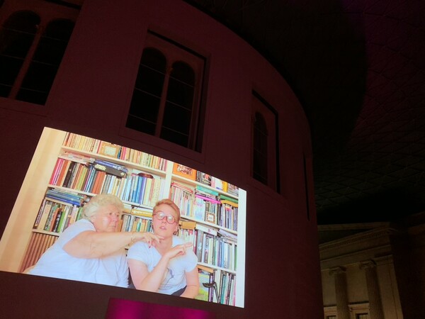 A large projector is showing two women, one older and the other younger. The older one has her hand under the others chin