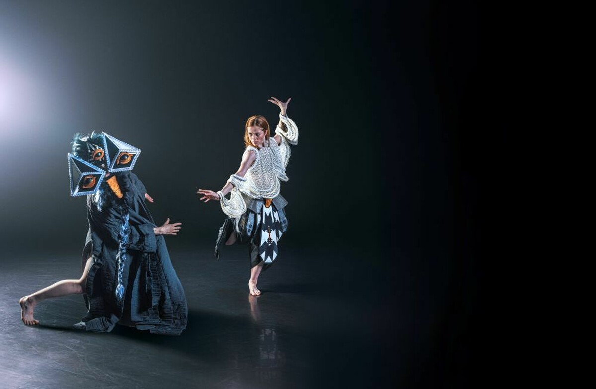 A person wearing a fantastcial costume with a three large eyes dancing on stage next a woman wearing a white beaded costume.