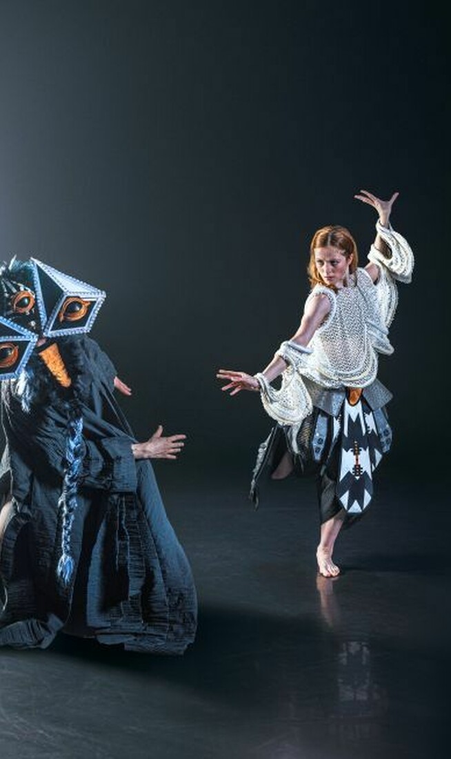 A person wearing a fantastcial costume with a three large eyes dancing on stage next a woman wearing a white beaded costume.