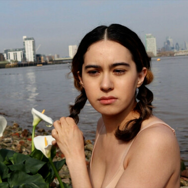 Medium close-up of a young woman. She is wearing a nude bra, braiding her dark hair, and she is sitting next to white lilies on a river bank. The background is the London Thames river, and skyscrapers.