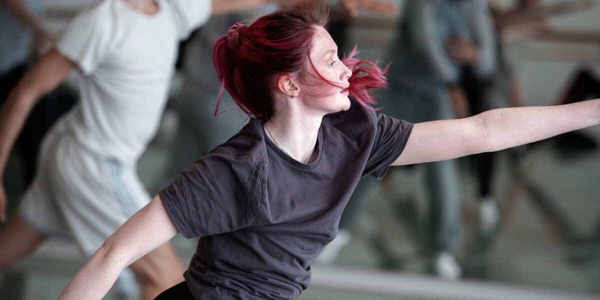 Several dancers are in the same position in one of our studios. The dancer in the foreground has pink hair and wears a dark grey t-shirt and black bottoms. They a lunging forward in a slight twist, looking towards their left arm which is fully stretched out in front of them, parallel to the floor. The other arm is lifted away from the body and is directed towards the floor.