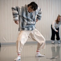 In the studio, a dancer in black bottoms and a light grey t-shirt sits upright with both knees bent and their forearms wrapped around their knees. They are looking at a dancer in the foreground who is wearing white trousers and a light striped hooded top. Their knees are bent and their arms are bent at a right angle towards the floor. They are looking down. Behind and to the right of them, a final dancer in a white top and black bottoms leans forward and holds their arms away from their body with elbows bent.