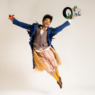 A performer jumps with both arms raised, wearing a blue tailcoat and yellow trousers and socks