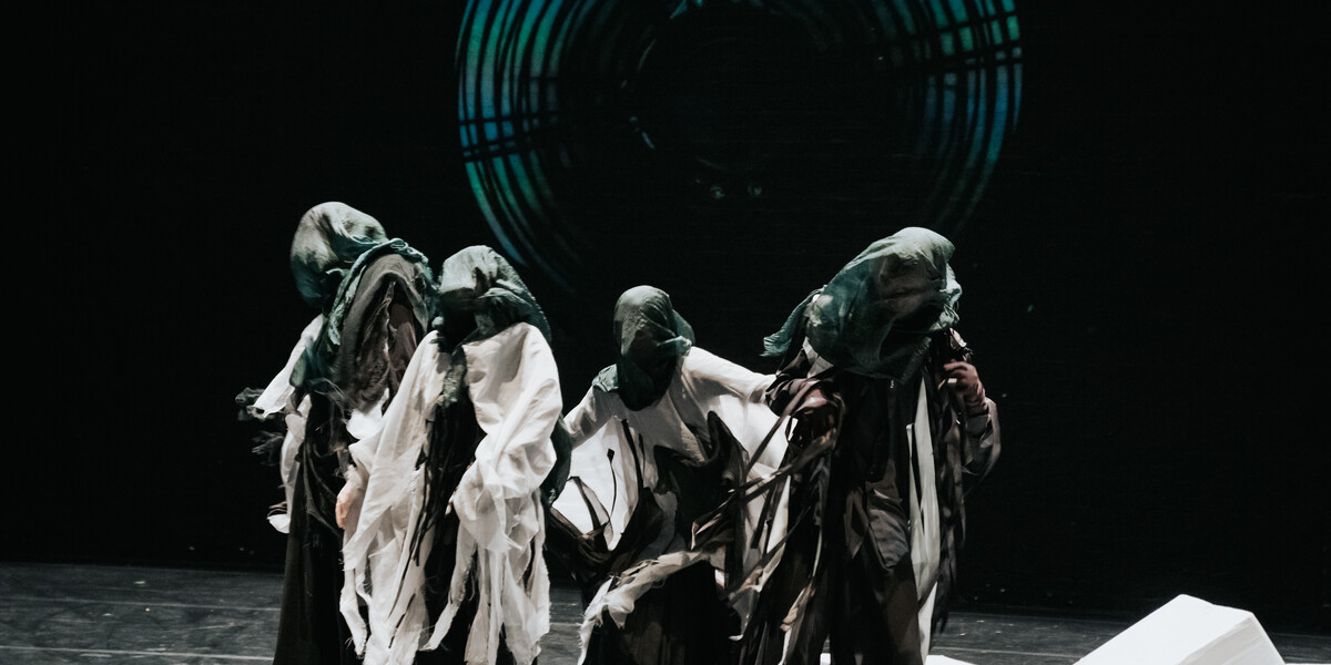 Four dancers are centre stage. They heads are covered in a dark green material and they are wearing grey, white and black baggy clothes, with tassels coming off them. There are white square boxes across the floor and a projection in the background of a fan.